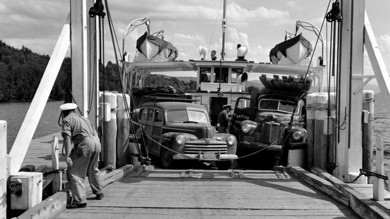 Historic black and white photo of ferry loaded with vintage cars and trucks at dock.