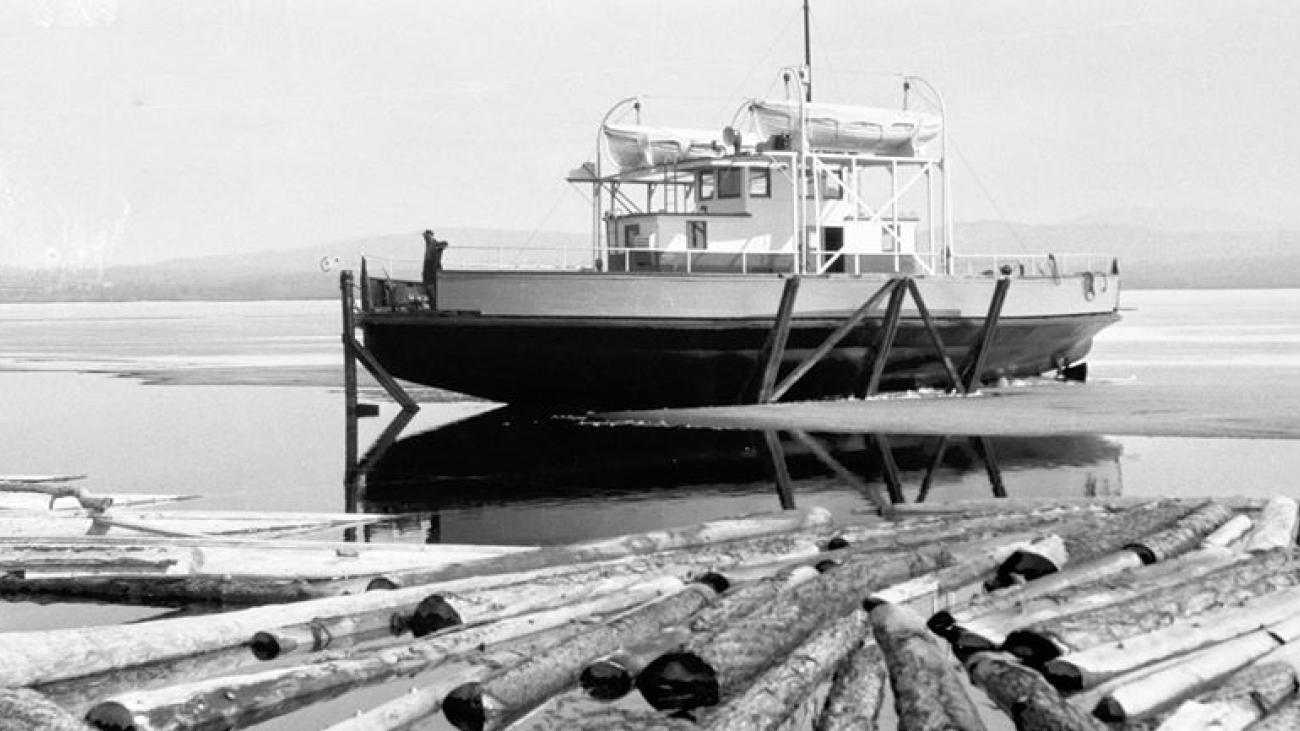 Boat docked by calm water, logs floating in foreground.