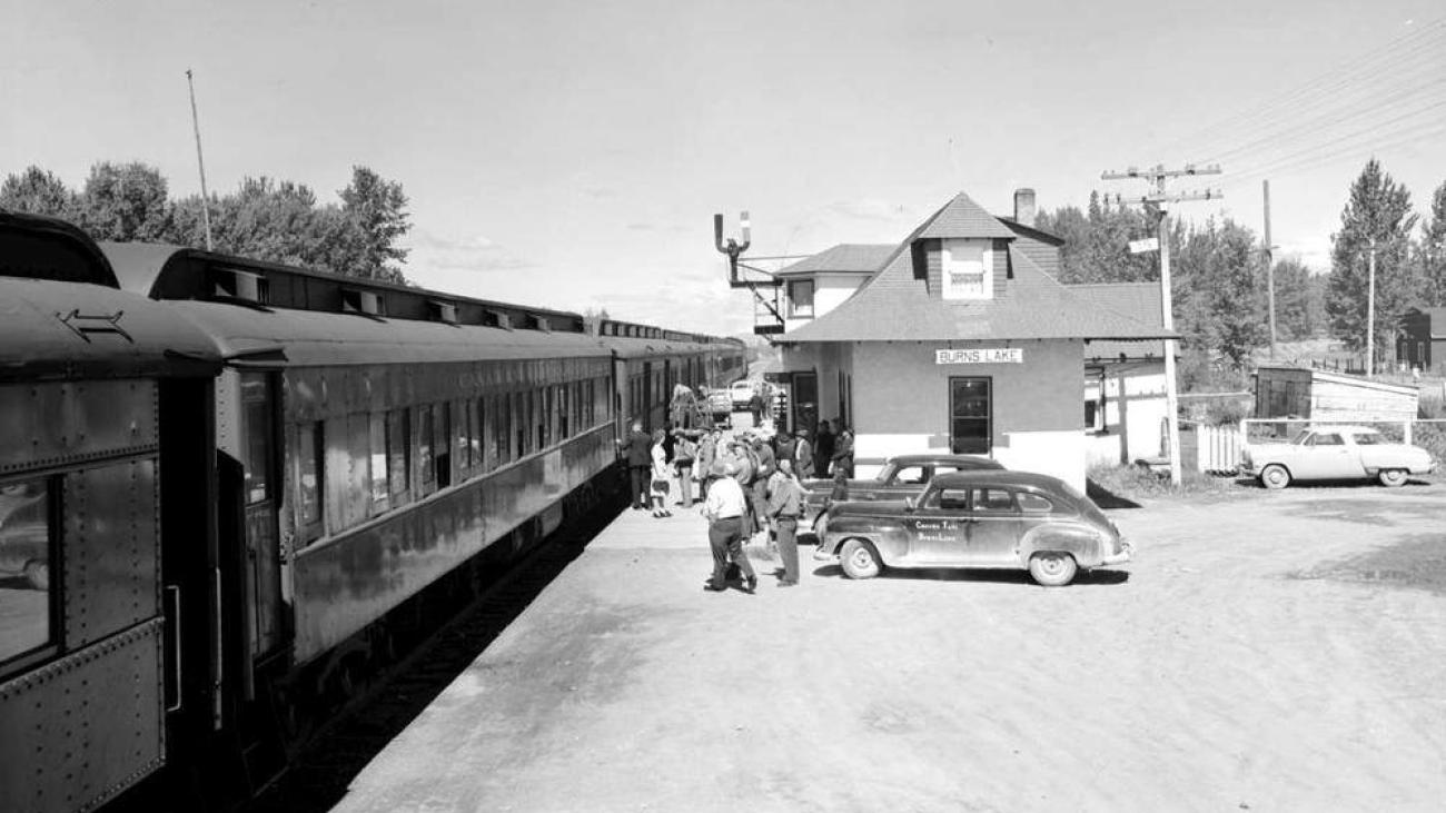 Train at a small station, vintage cars parked nearby, and people standing on the platform.