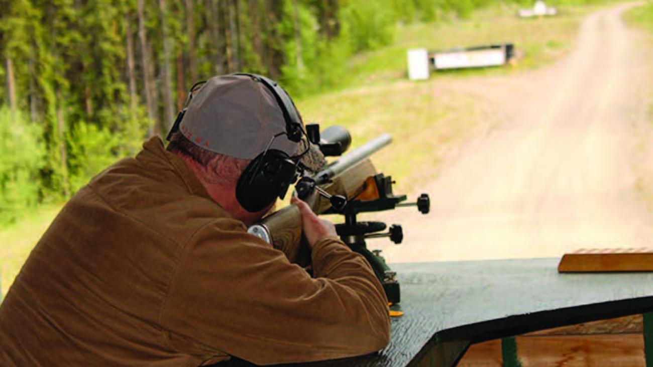 Man aiming a rifle at a shooting range, wearing headphones and cap.