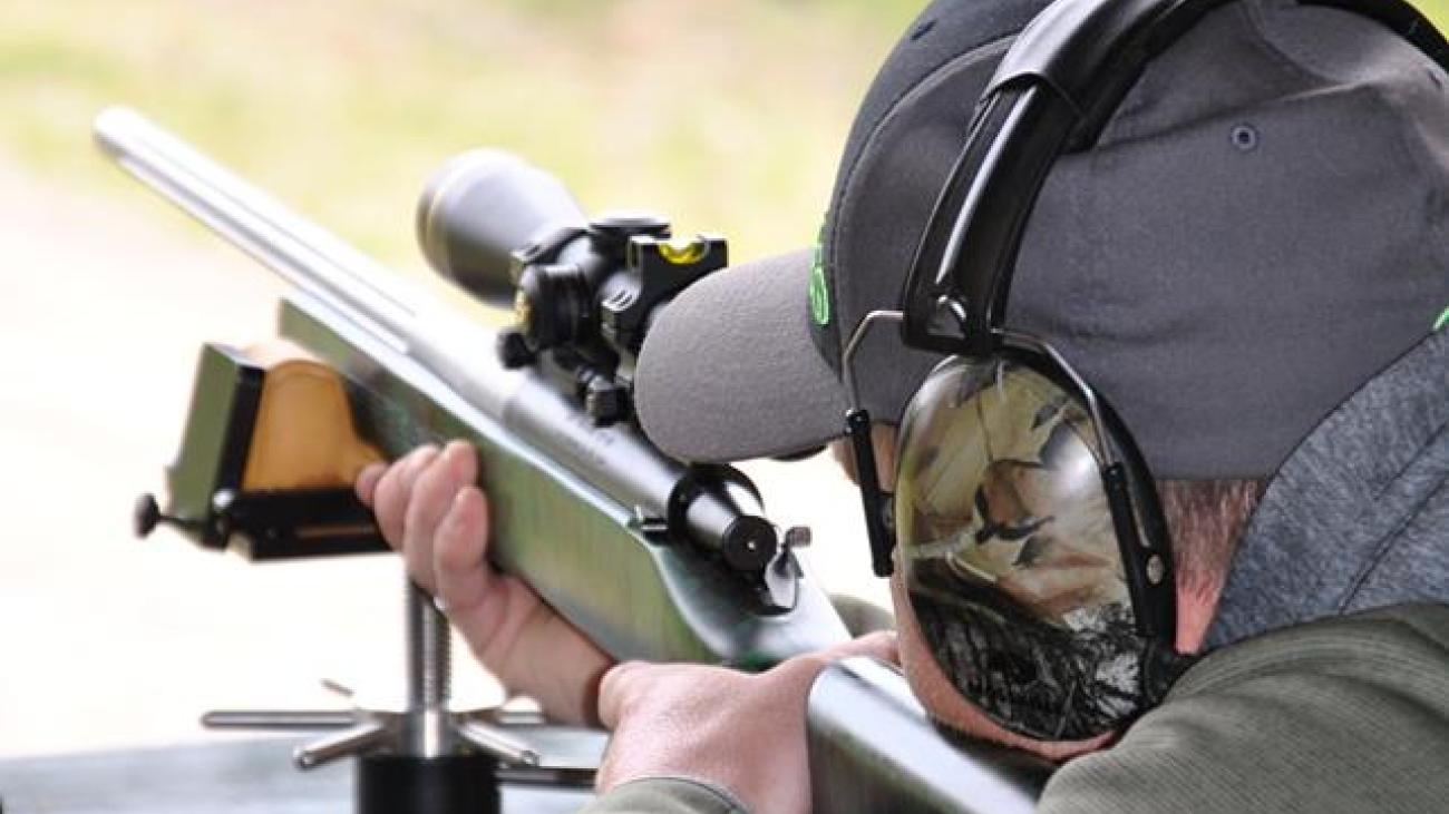 Man aiming rifle at outdoor shooting range, wearing ear protection.