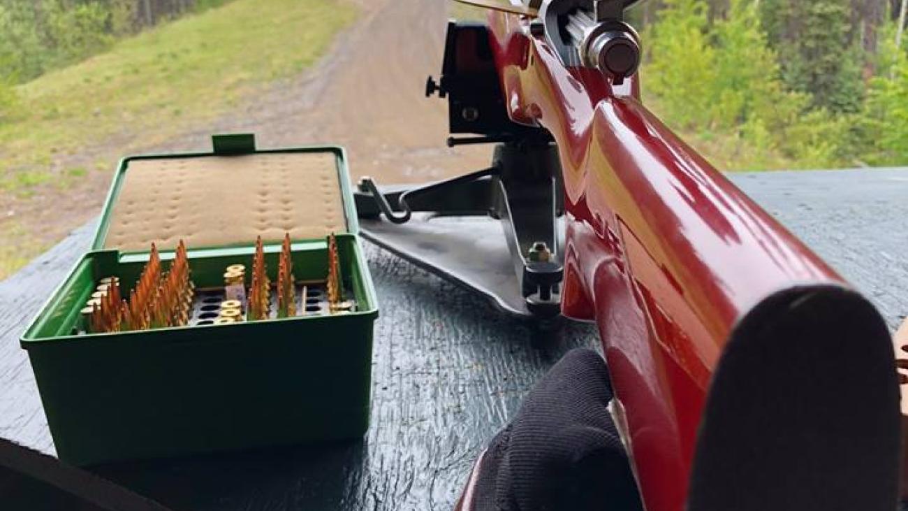 Wooden rifle with scope on a table, ammunition box beside it, forest in background.