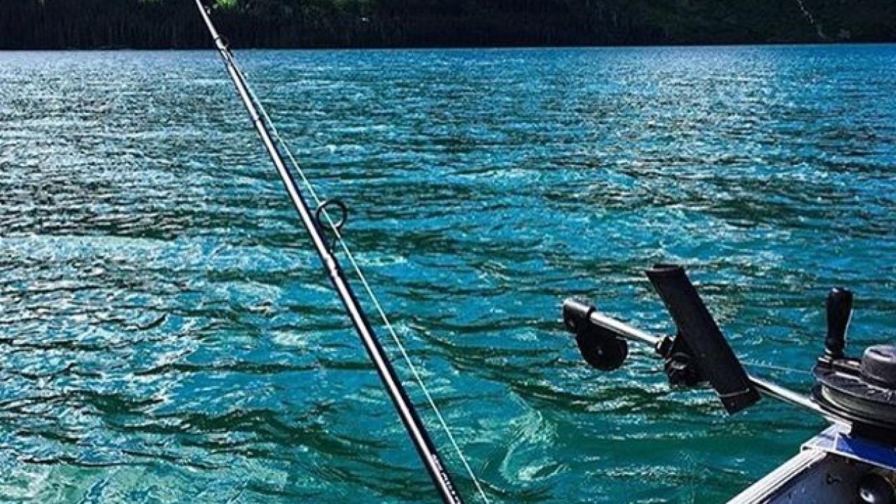 Fishing rod on a boat, calm blue lake with forested mountain backdrop.