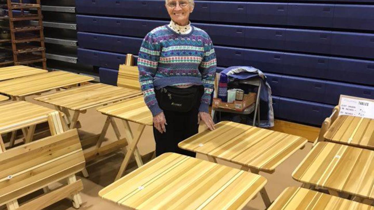 Elderly person stands among several wooden folding tables in a gym.