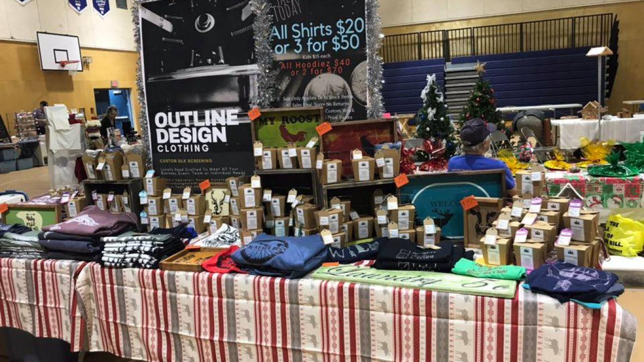 Craft fair table with t-shirts, candles, and holiday decorations.