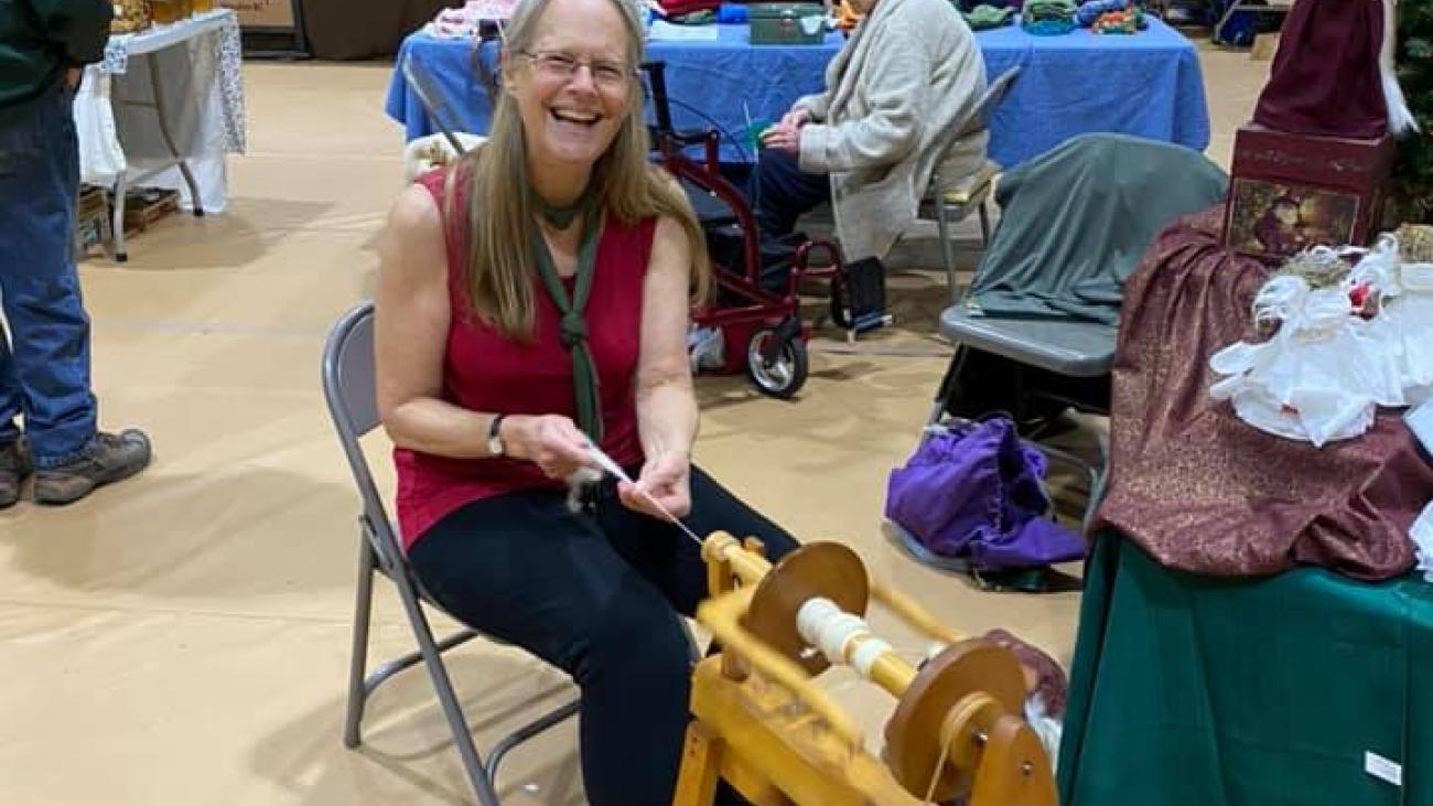 Woman spinning yarn at a fair, surrounded by tables and people.