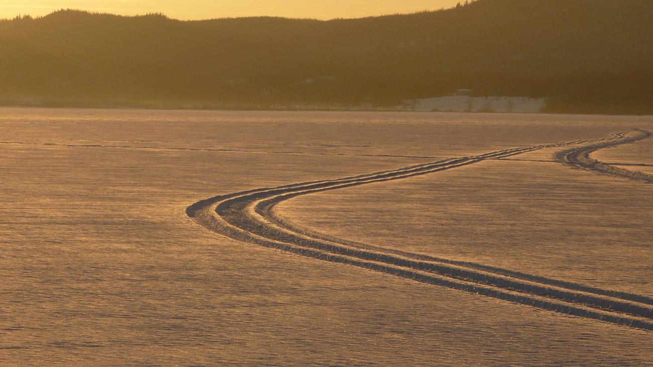 Snowmobile or vehicle tracks curving across a frozen lake at golden hour.