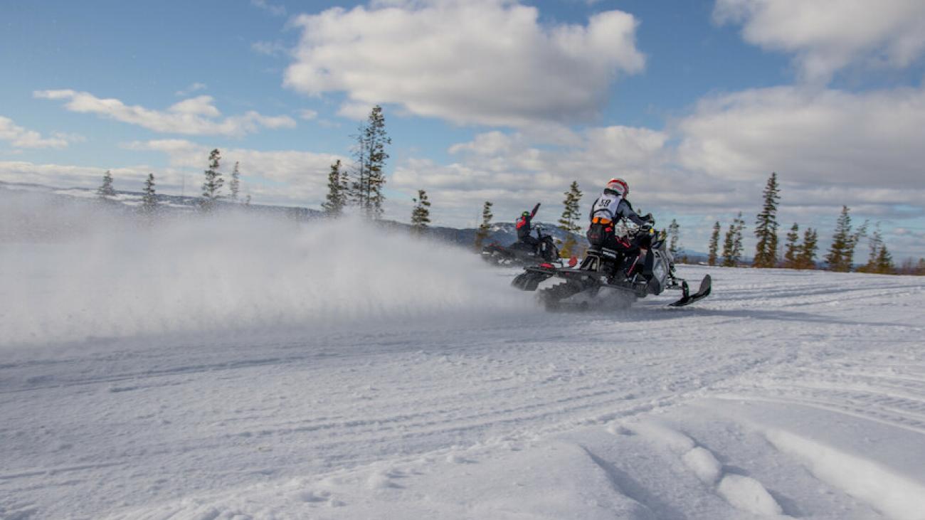 Snowmobiles racing on a snowy field under a cloudy blue sky.