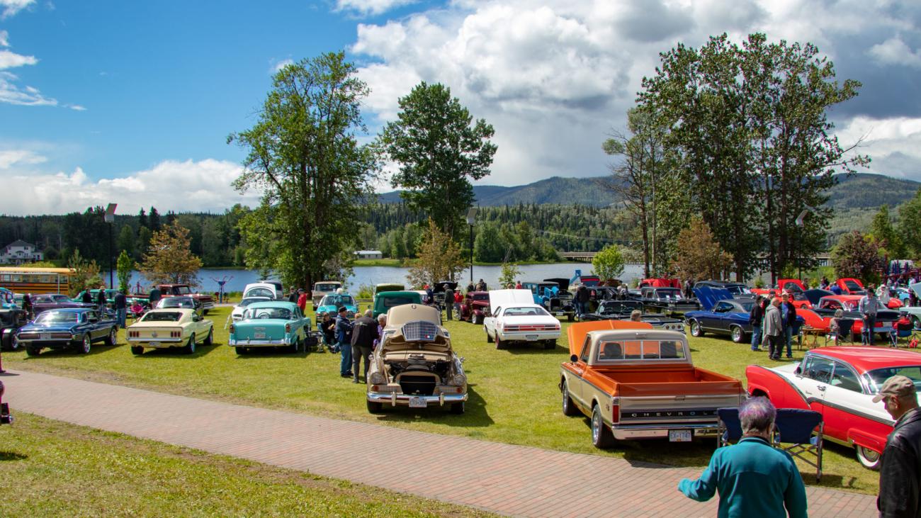 Classic cars displayed on a grassy field by a lake, with trees and mountains in the background.