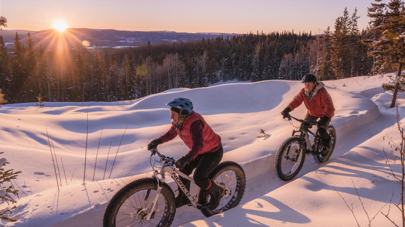 Two cyclists ride fat bikes on a snowy trail at sunset.