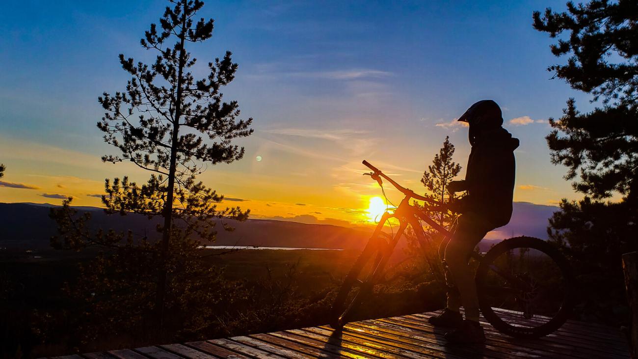 Cyclist silhouetted against a sunset, standing with a bike on a forested overlook.