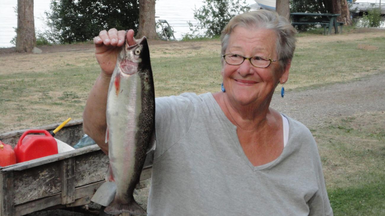 Smiling woman holding a trout beside a lake.