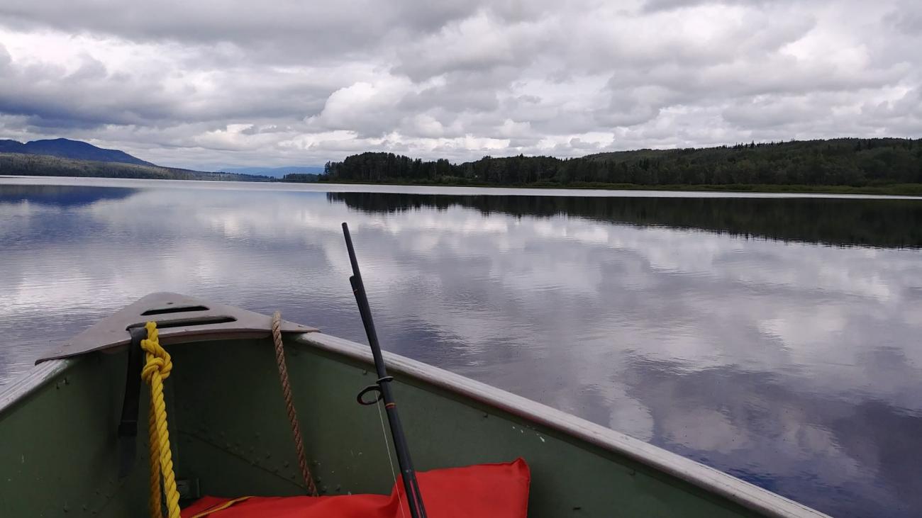 Small boat on a calm lake with fishing poles; cloudy sky reflects on water.