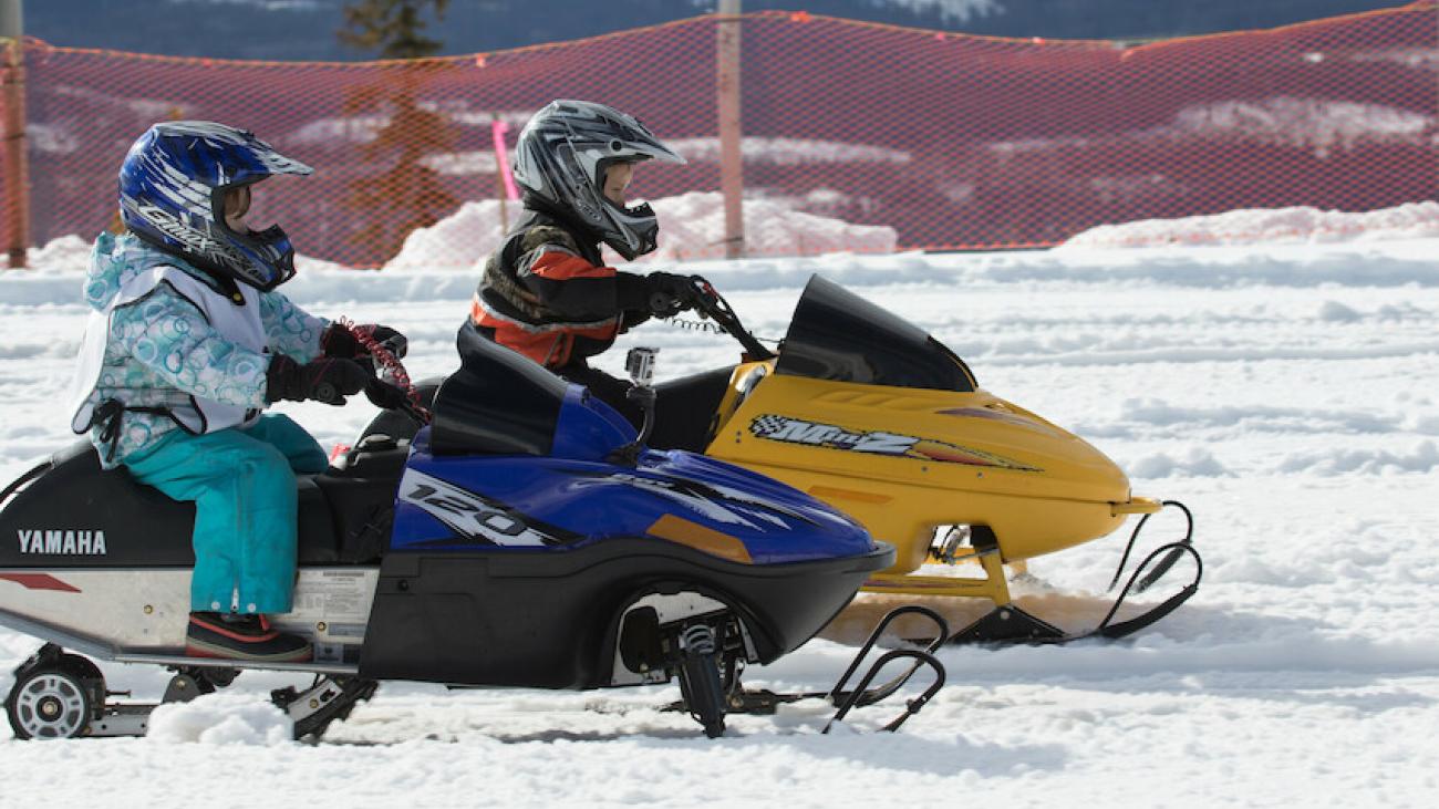 Two children racing on mini snowmobiles in snowy landscape.