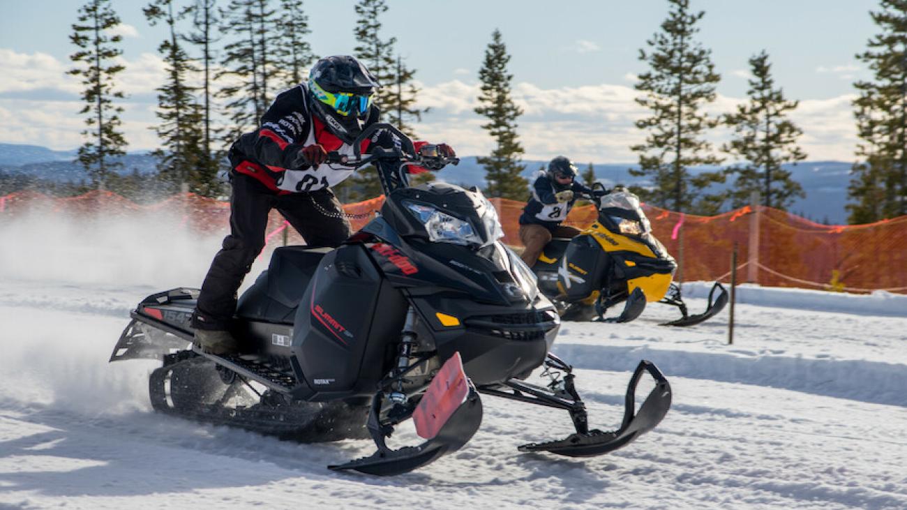 Two people racing snowmobiles on a snowy track.