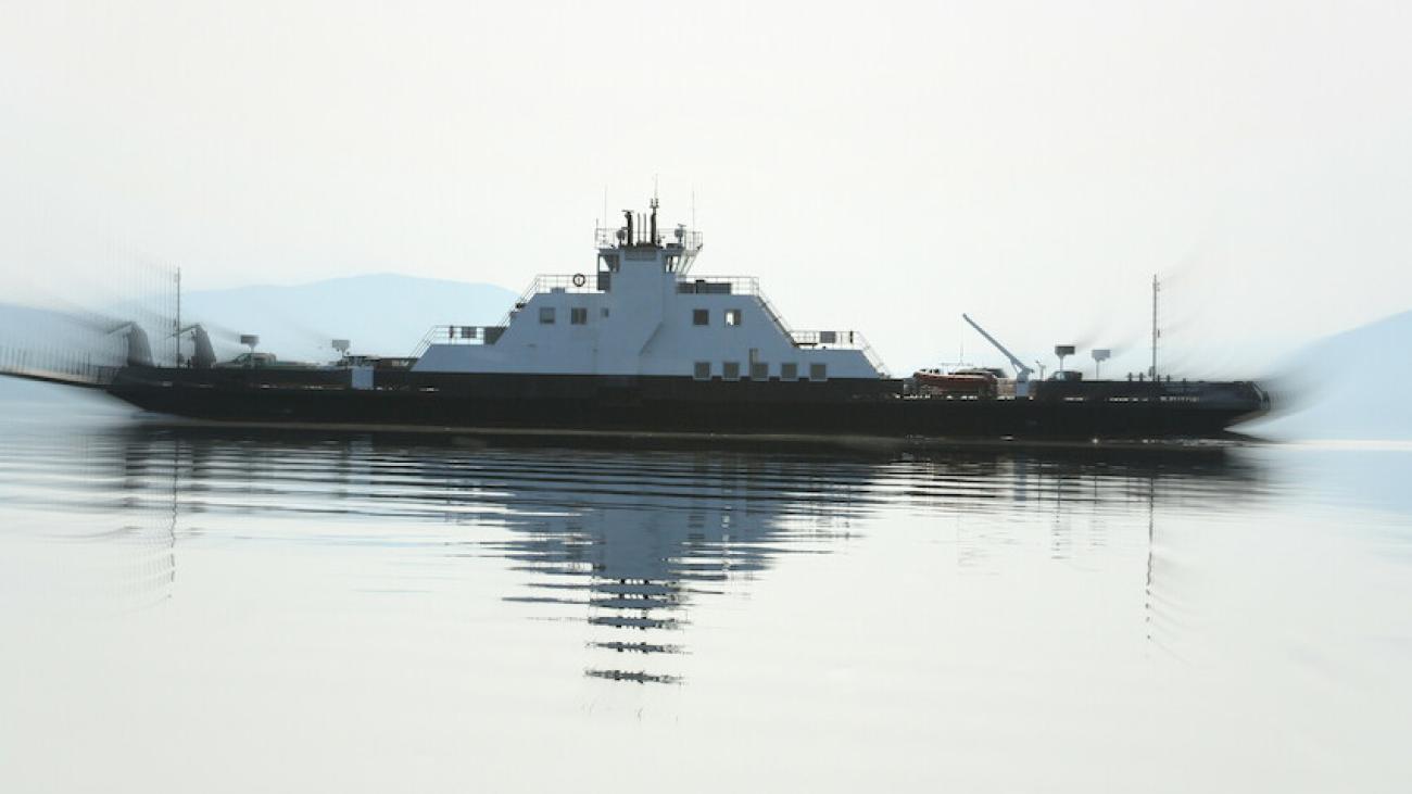 A ferry reflected on calm water against a hazy, mountainous backdrop.