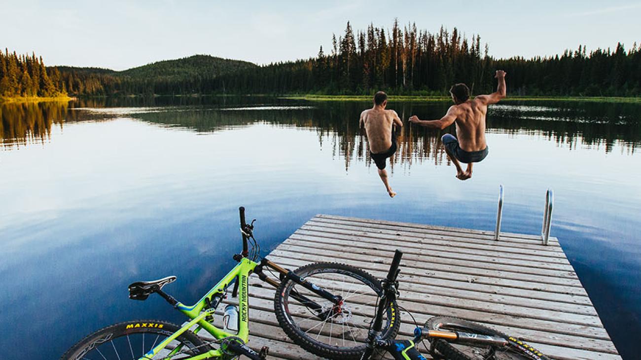 Bicycles on a dock, two people jump into a calm lake surrounded by trees.