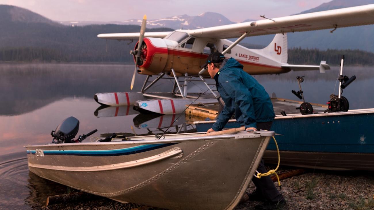 Man in blue jacket with a boat near a floatplane on a lake at dawn.