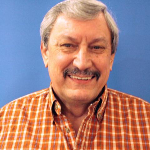 Smiling man with gray hair and moustache, wearing a plaid shirt against a blue background.
