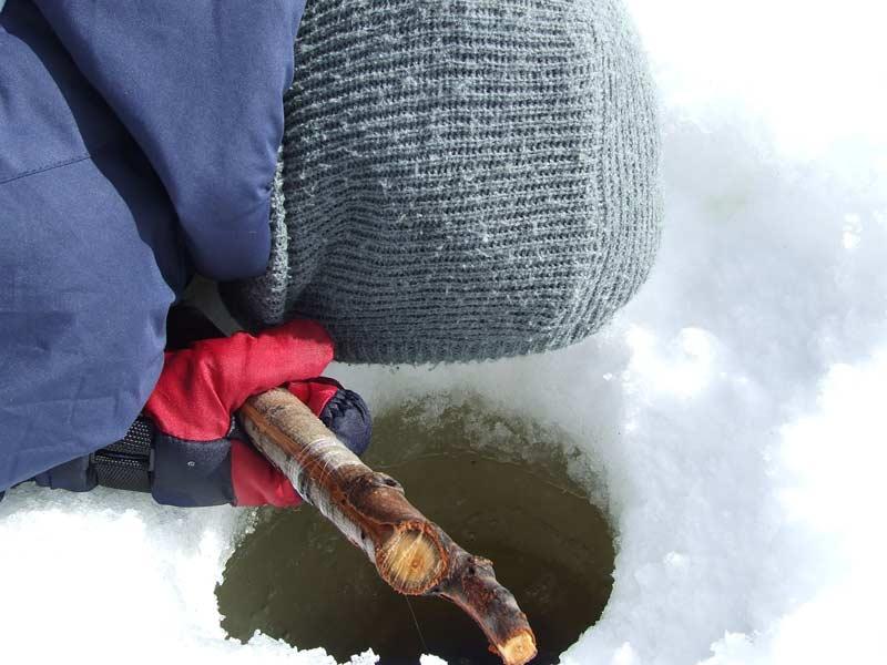 Person peering into ice hole, holding stick, dressed warmly in gray and blue.